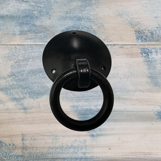 Black forged iron barn dome cabinet pull with a circular ring and rustic finish, mounted on a light wooden background.
