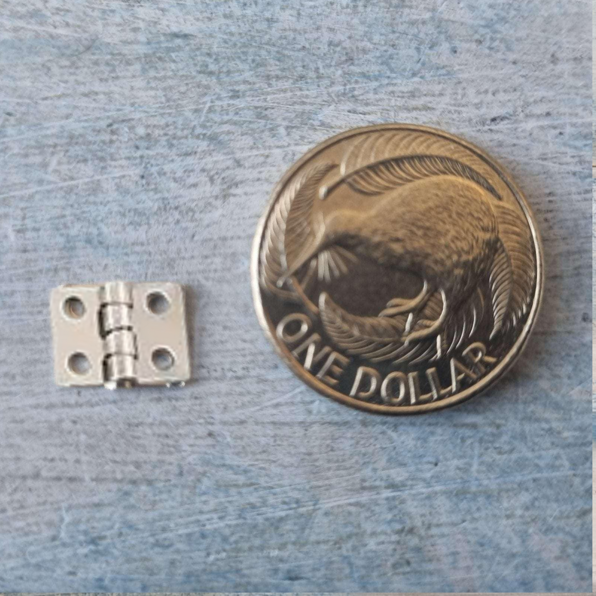 A small silver metal hinge shown next to a one dollar coin for scale, on a textured blue-gray surface.