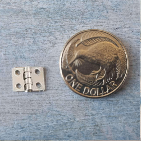 A small silver metal hinge shown next to a one dollar coin for scale, on a textured blue-gray surface.